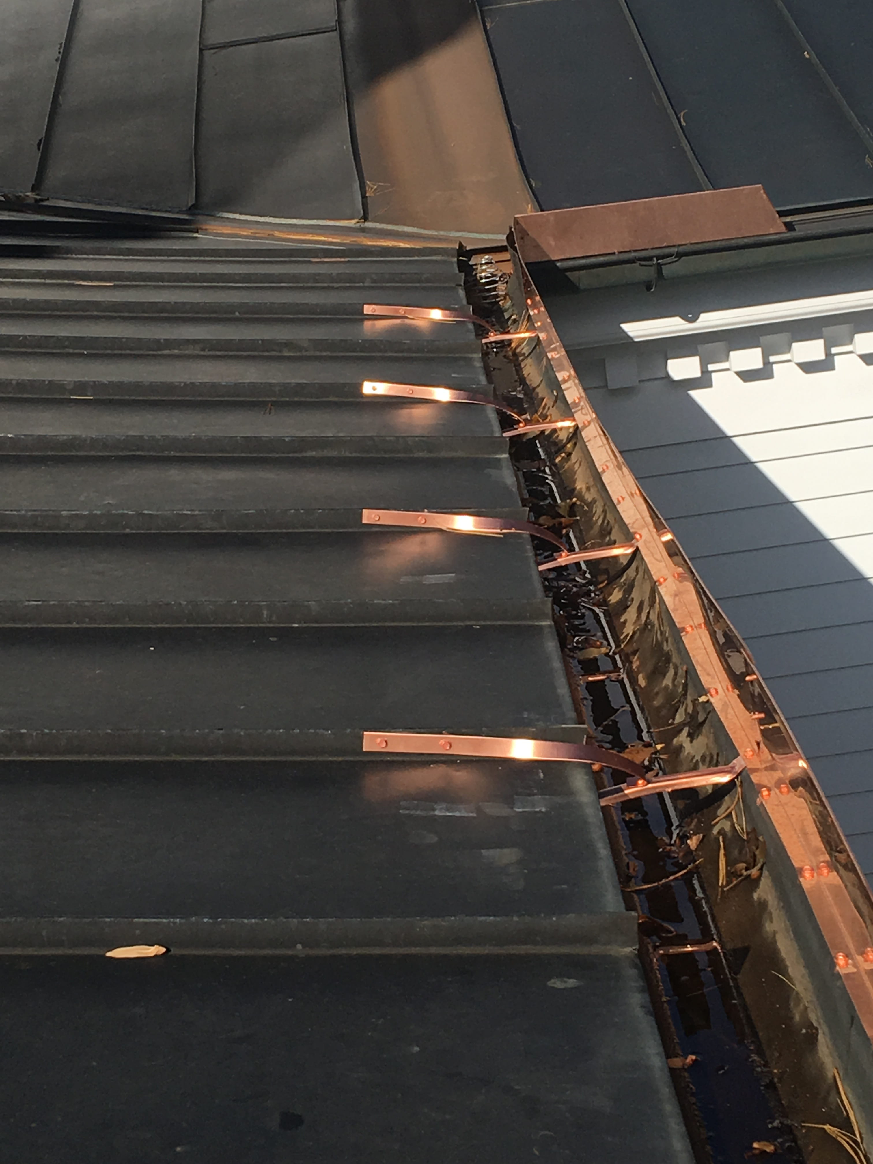 Copper gutter with exposed hanger brackets and standing water from organic debris buildup on metal roof